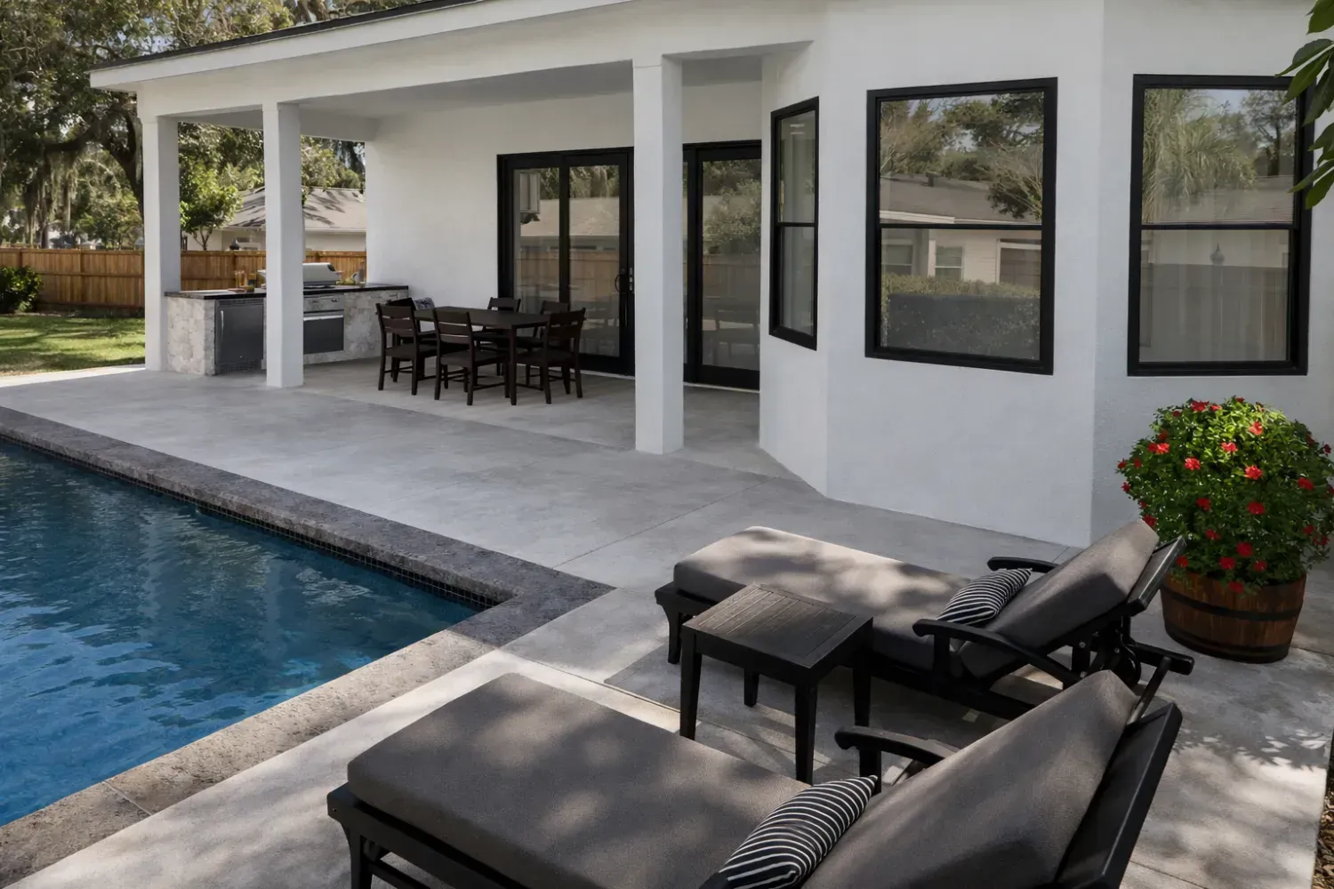A white house with a covered outdoor patio, dining area, swimming pool, and lounge chairs on a paved deck.