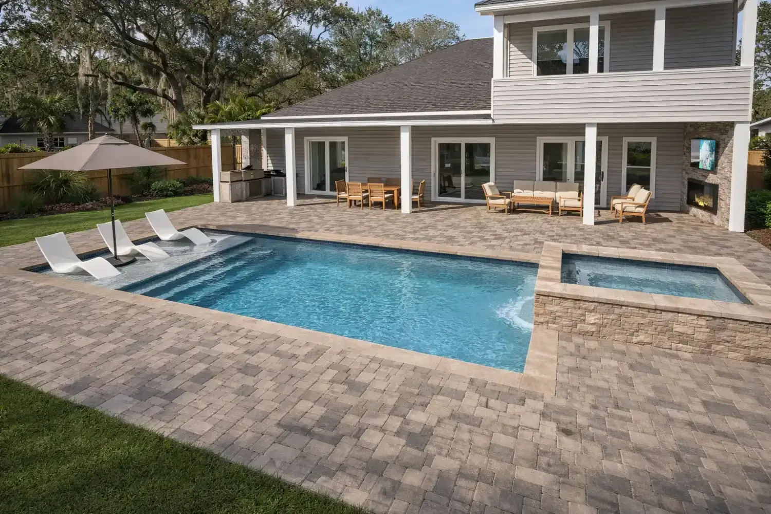 A backyard features a rectangular swimming pool, a raised spa, stone pavers, white lounge chairs, and a covered patio.