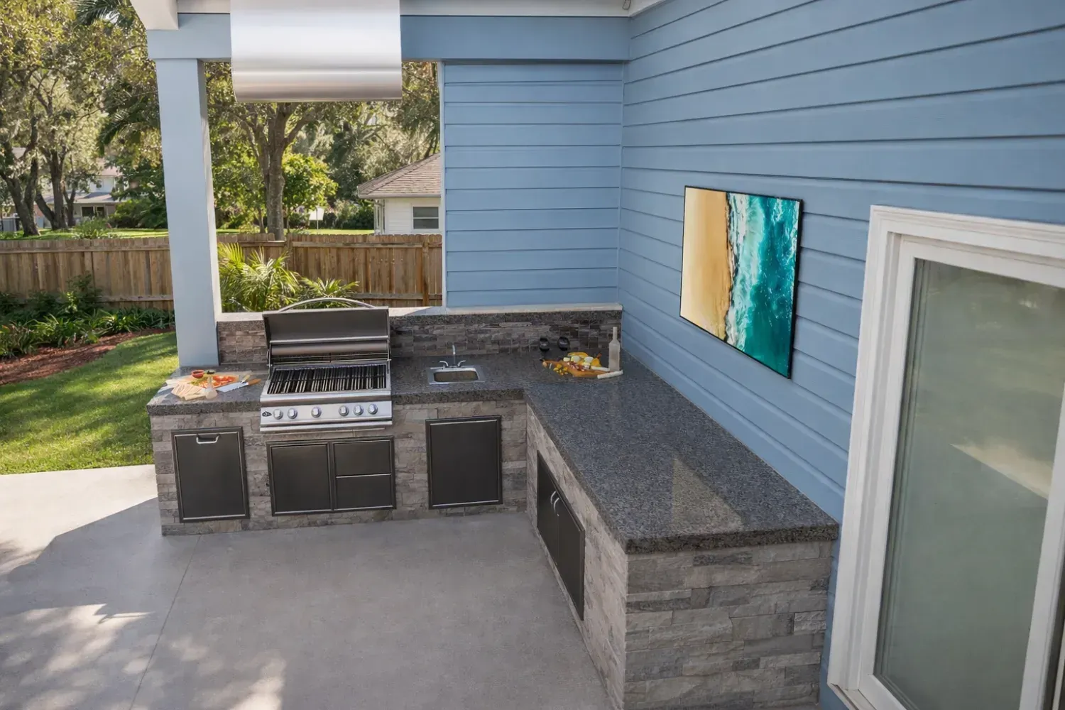 An L-shaped outdoor kitchen with a grill, stone base, and granite countertop, situated on a patio against a blue wall.