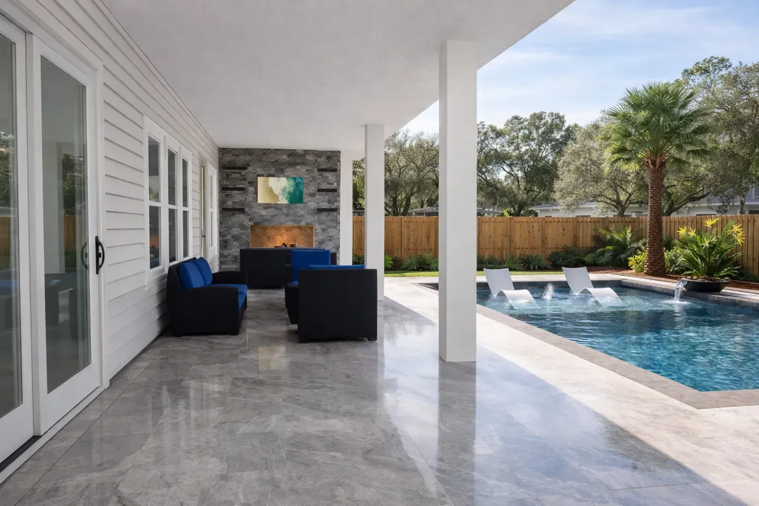 Covered patio with a stone fireplace, dark seating, and a swimming pool with lounge chairs in a backyard.