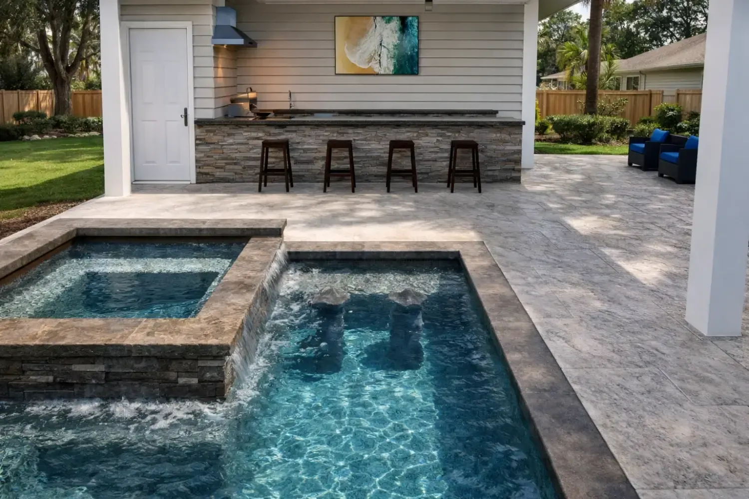 Outdoor patio featuring a rectangular pool and attached hot tub, with a stone-accented bar and four stools in the background.