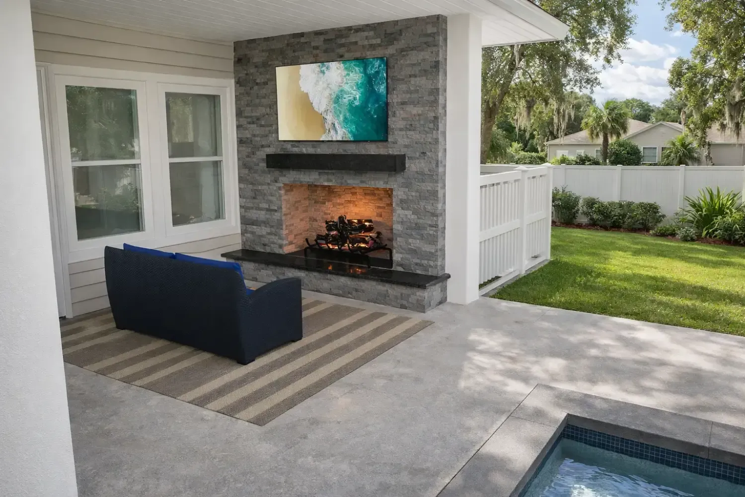 A stone fireplace and TV on a covered patio with a blue sofa on a striped rug, next to a small pool and grassy backyard.