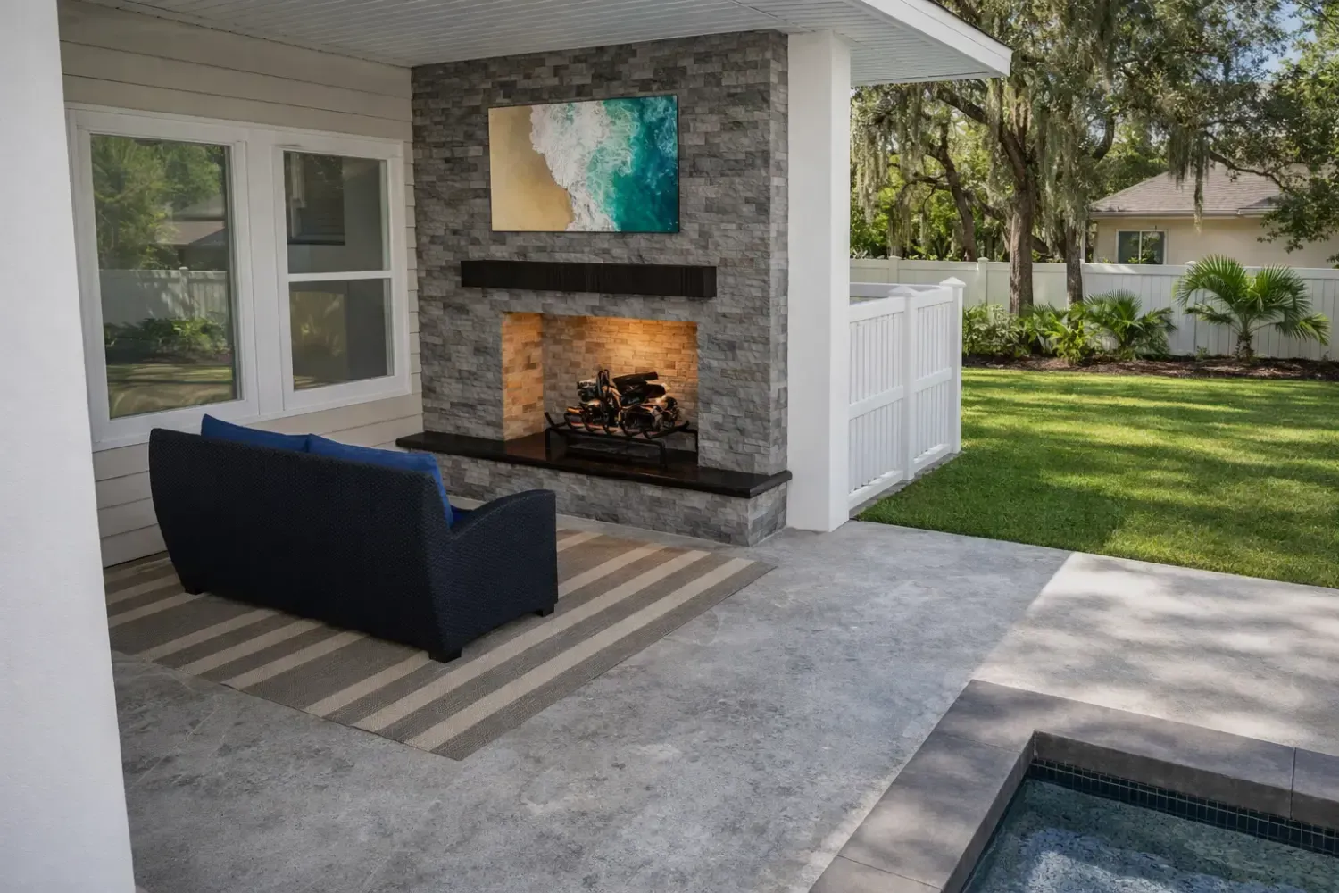 A cozy outdoor patio with a stone fireplace, blue sofa, striped rug, and view of a lawn and pool area.