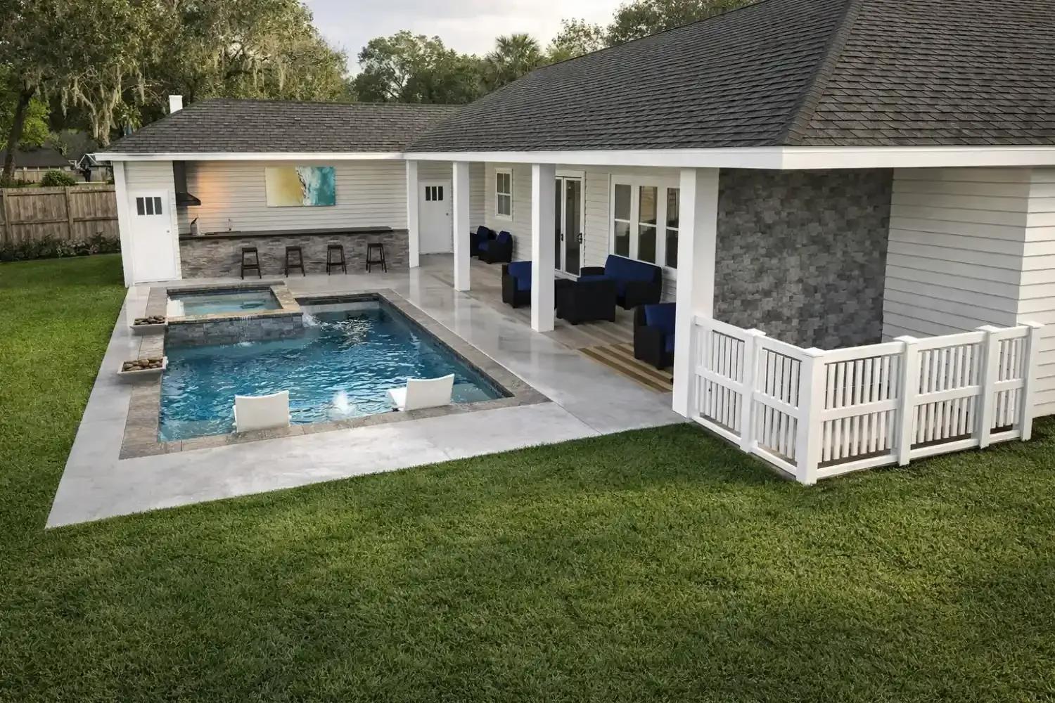 A backyard view of a home with a covered patio, outdoor bar area, swimming pool, and white picket fence on green grass.