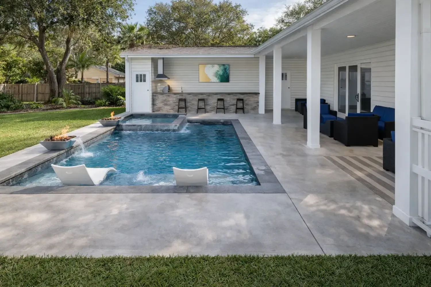 A swimming pool with two lounge chairs, a connected hot tub, and an outdoor bar area on a patio next to a white home.
