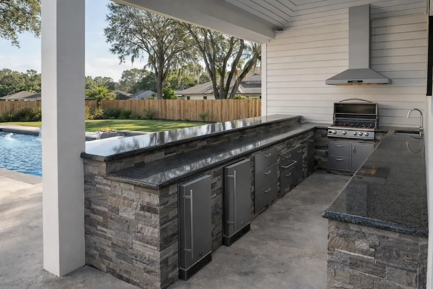 An outdoor kitchen featuring stone masonry, a granite bar top, built-in stainless steel appliances, and a grill hood.
