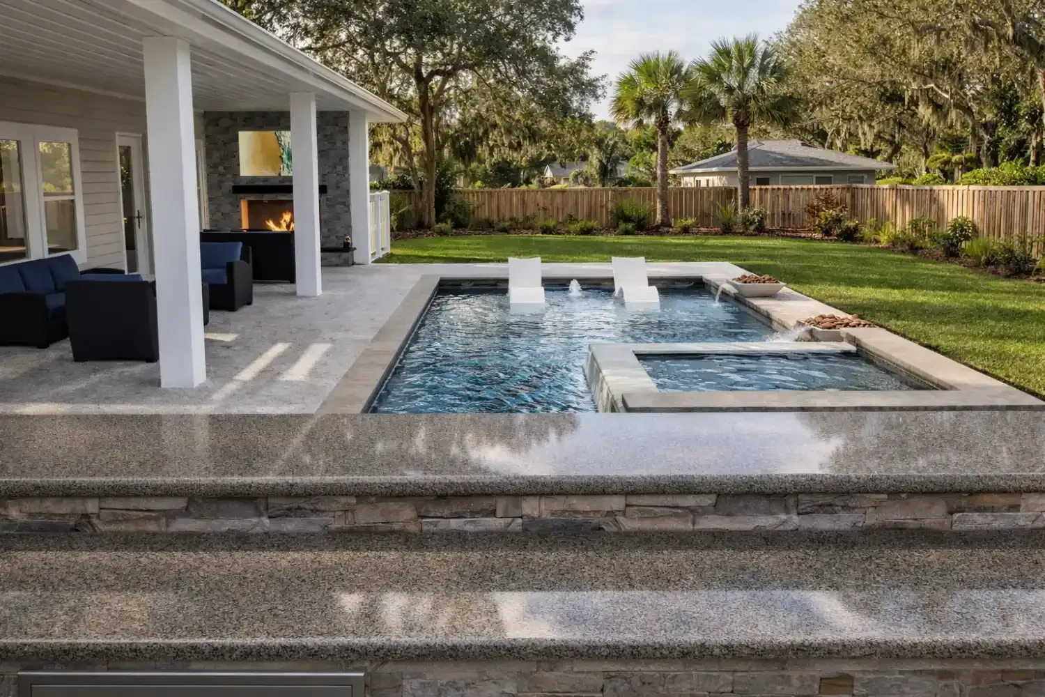 A covered patio with outdoor seating next to a swimming pool featuring tanning ledges and a stone fire pit.