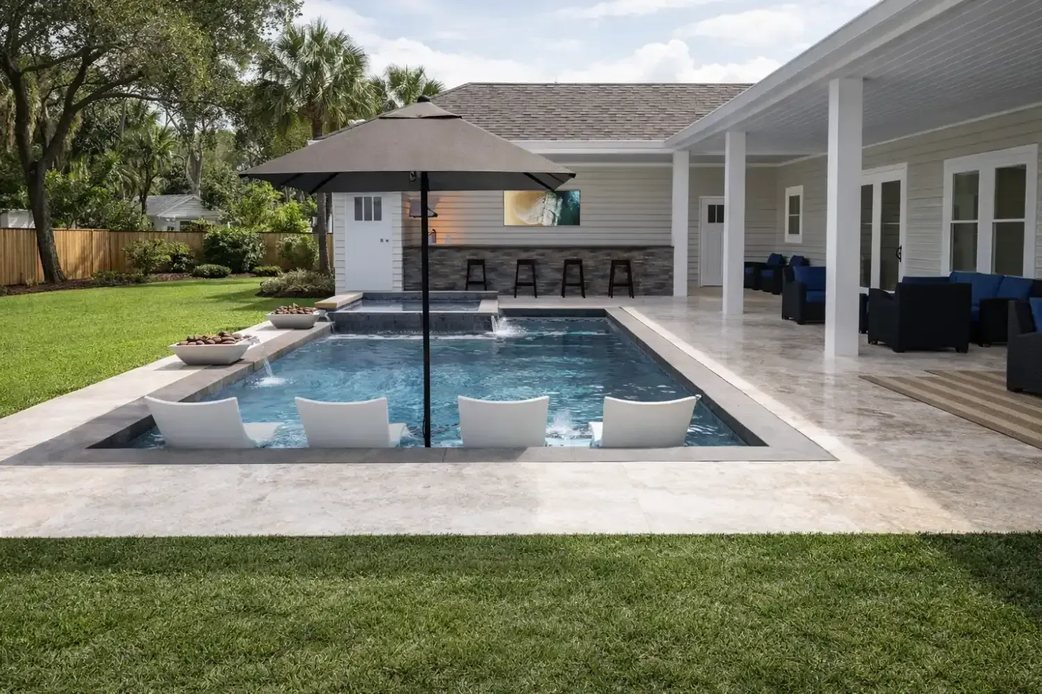 Backyard swimming pool with four white lounge chairs, an umbrella, and a covered patio seating area on a sunny day.