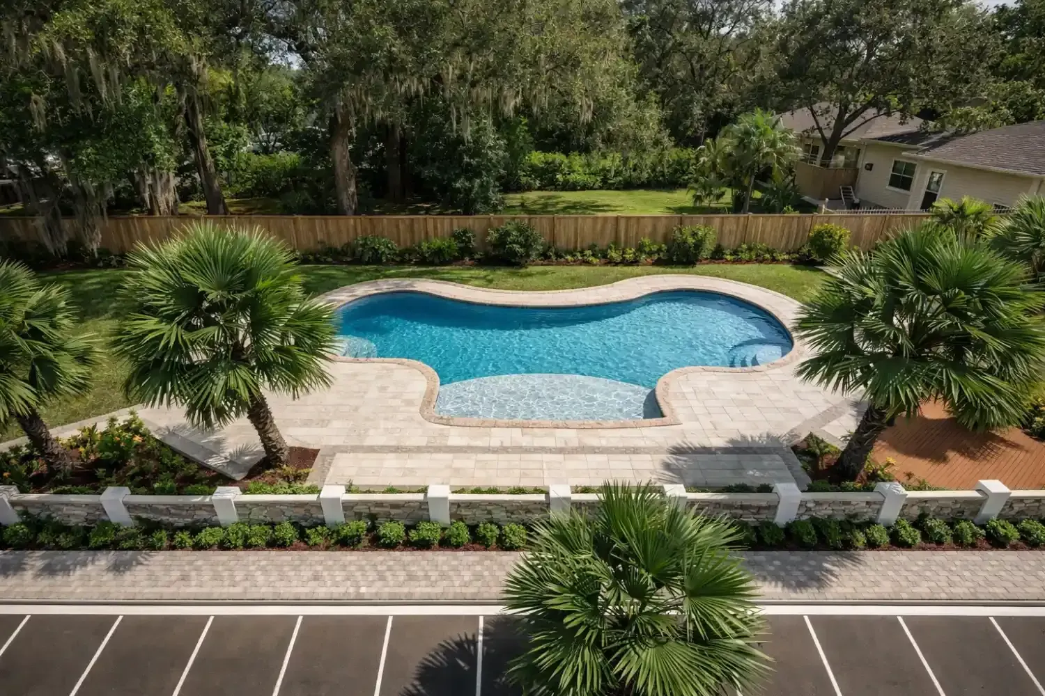 An outdoor kidney-shaped swimming pool with tiled steps, surrounded by pavers, palm trees, and a low white wall.