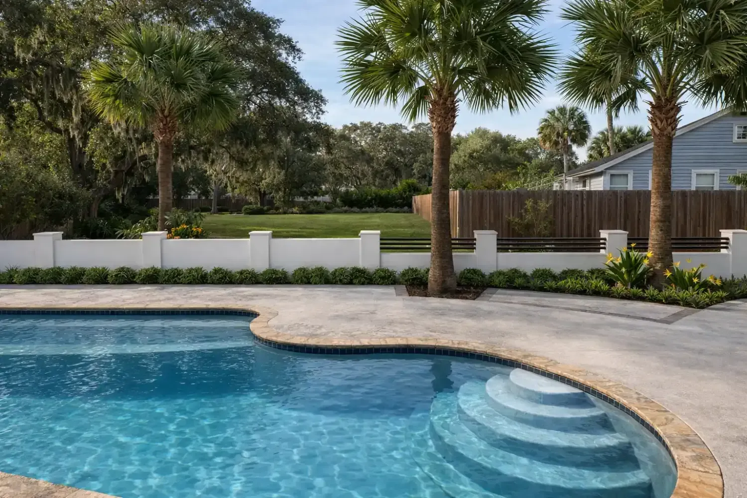 A sunlit backyard swimming pool with blue water and stone patio, featuring palm trees and a white wall against a blue sky.