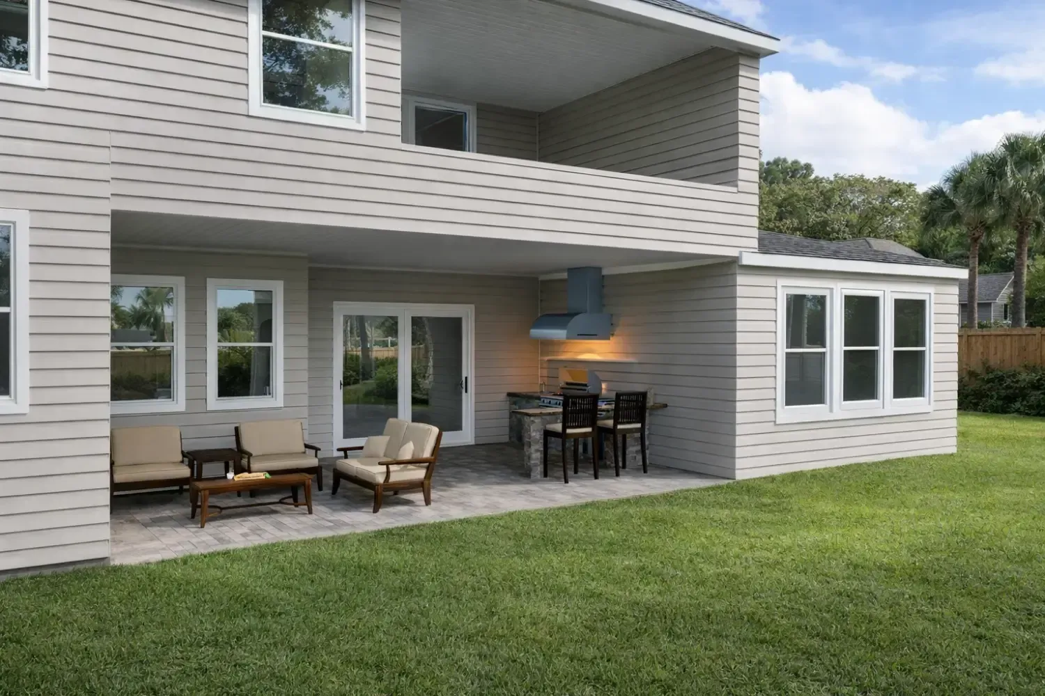 A two-story beige house with a covered outdoor patio, including a kitchen area, dining set, and seating on a green lawn.