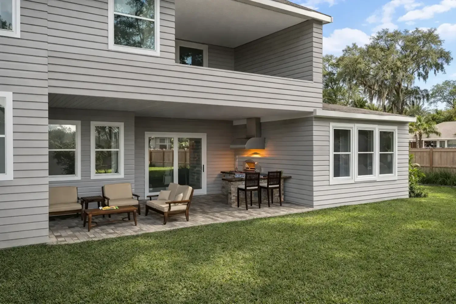 A grey house with a paved patio, outdoor furniture, and a small built-in grill, overlooking a green backyard.