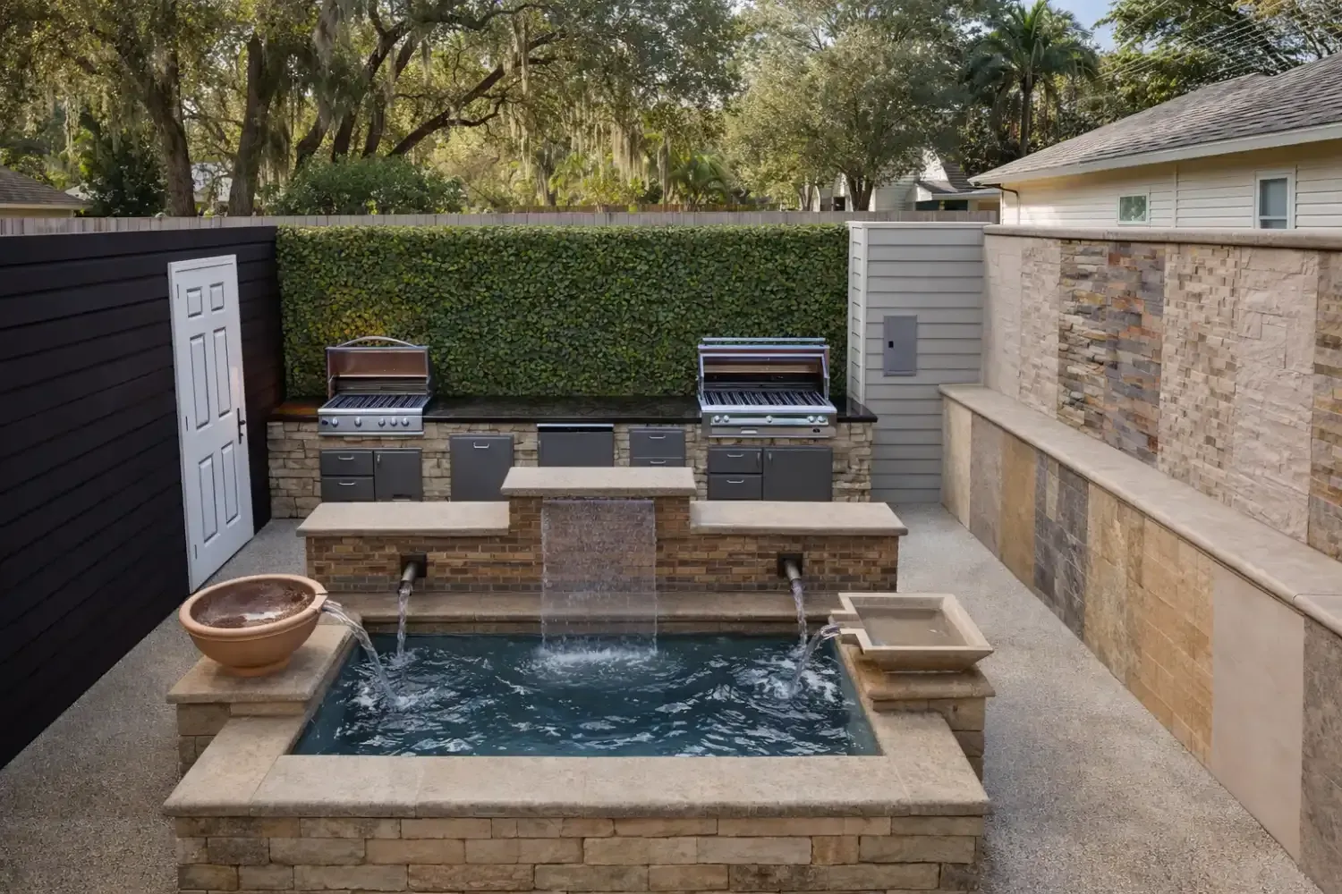 A stone patio features a rectangular plunge pool with a waterfall, fountain spouts, and an outdoor kitchen against a hedge.