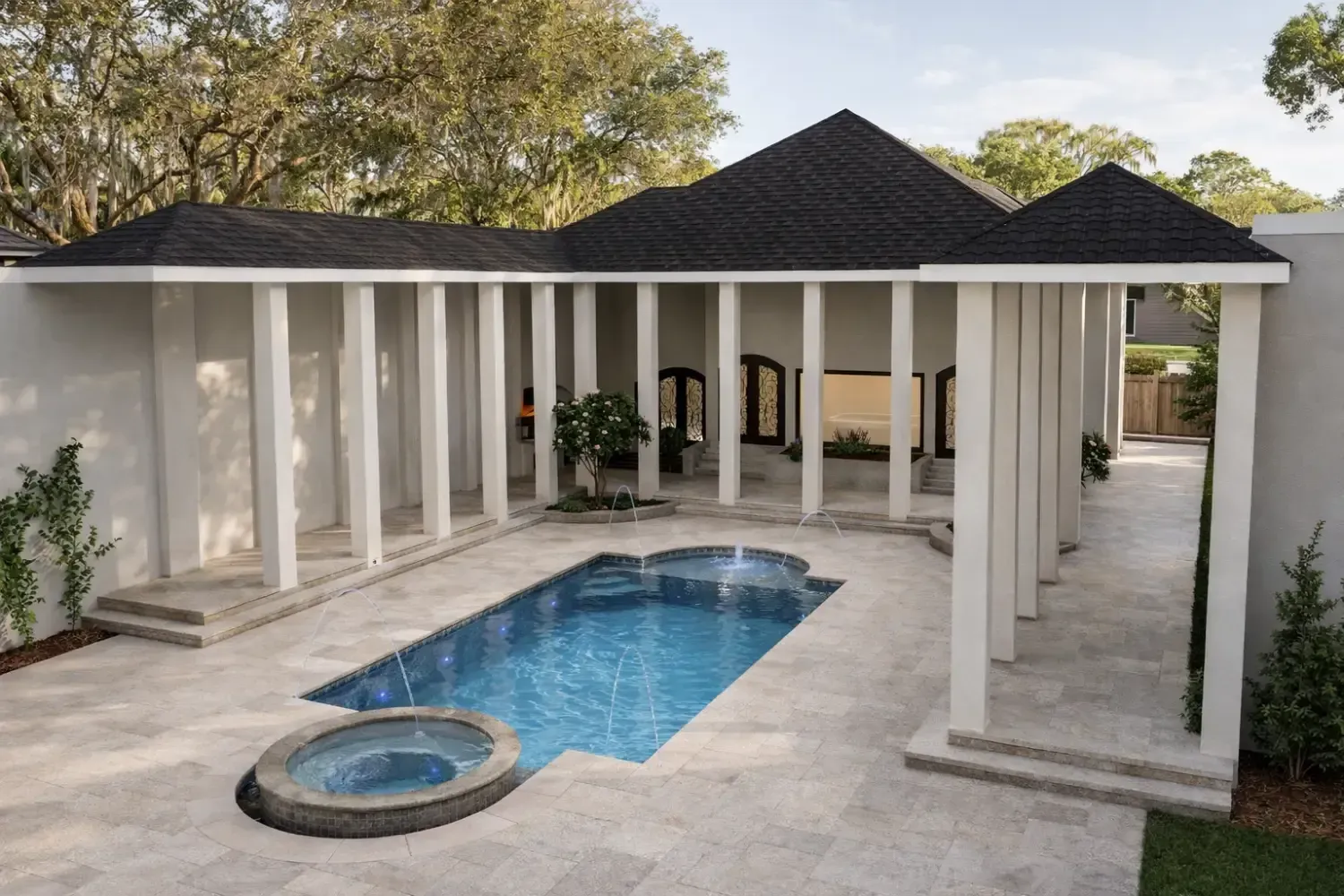 A rectangular pool and circular spa in a travertine courtyard bordered by a white house with a columned, covered patio.