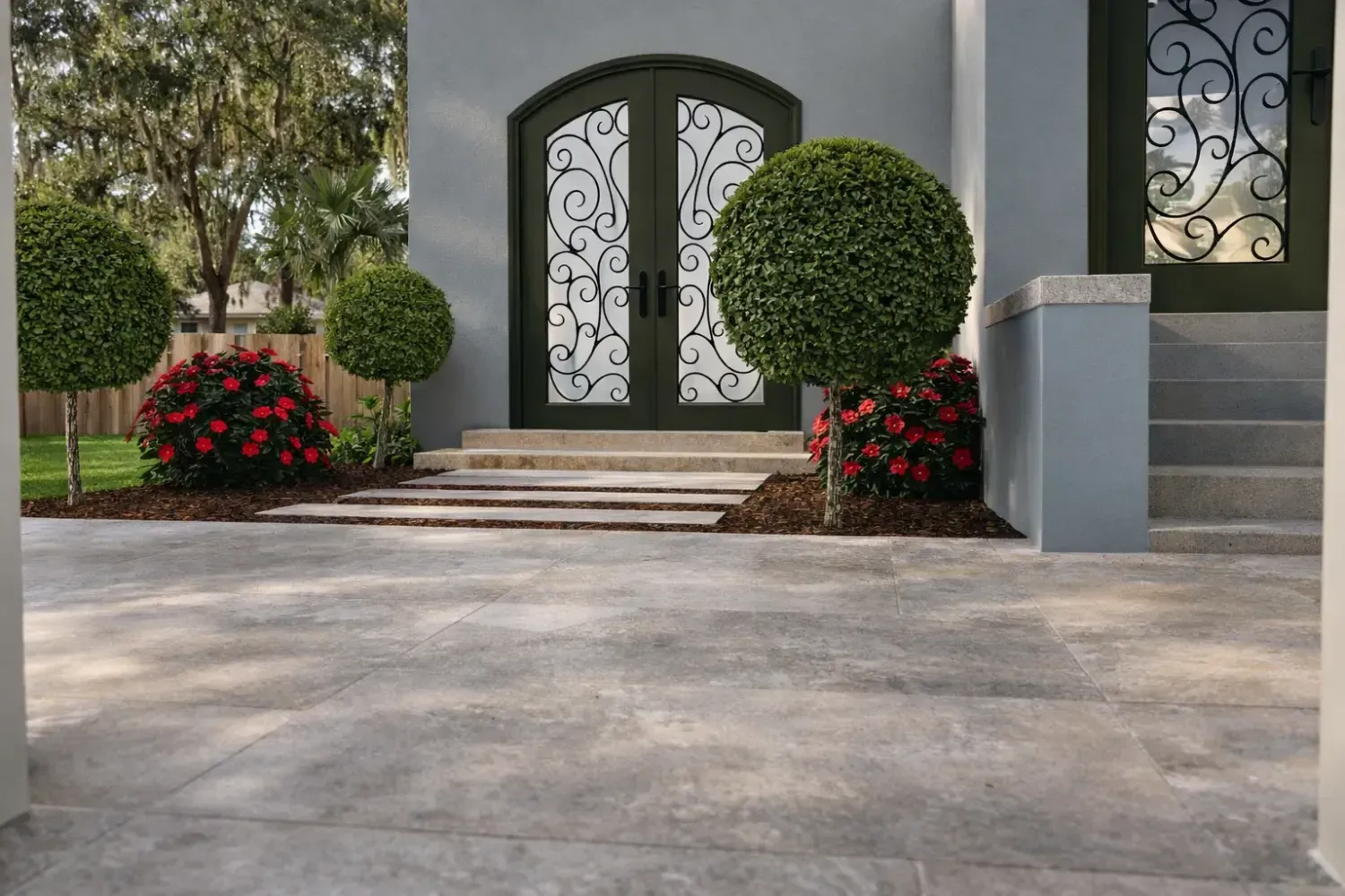 A paved patio entrance with topiary trees and red flowering shrubs flanking a dark, ornate arched double door.