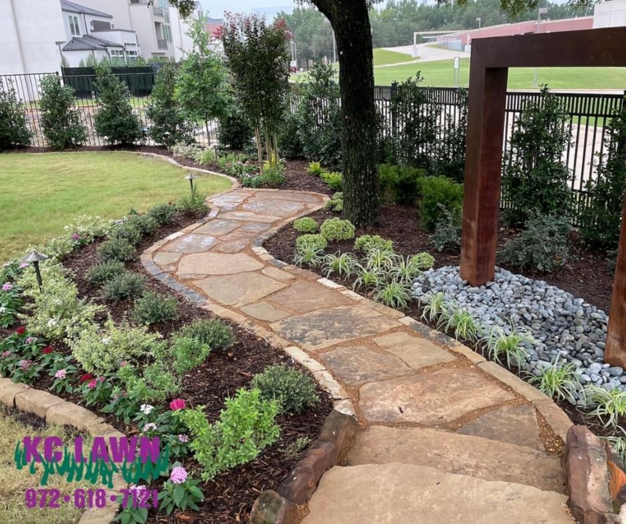 Stone pathway winds through a garden with flower beds and a tree.