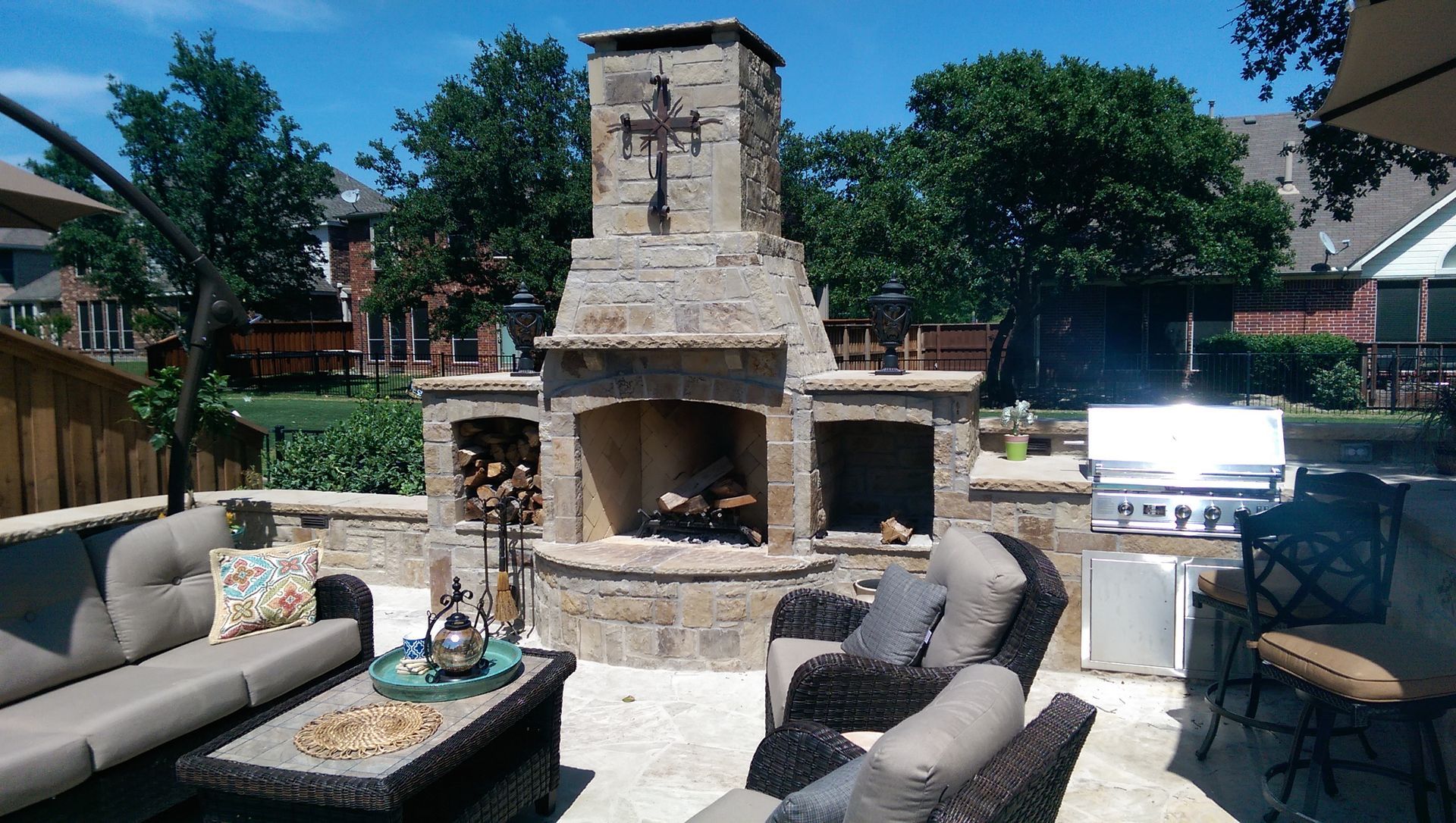 Stone outdoor fireplace with seating, grill, and wood storage, on a sunny patio.