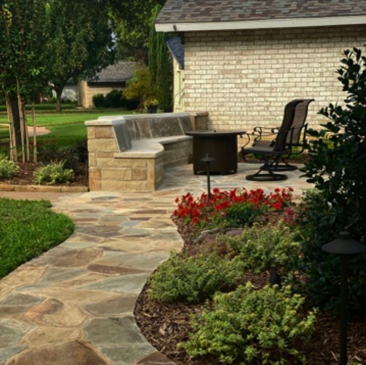 Stone pathway leads to patio with built-in bench, fire pit, and brick building with landscaping.