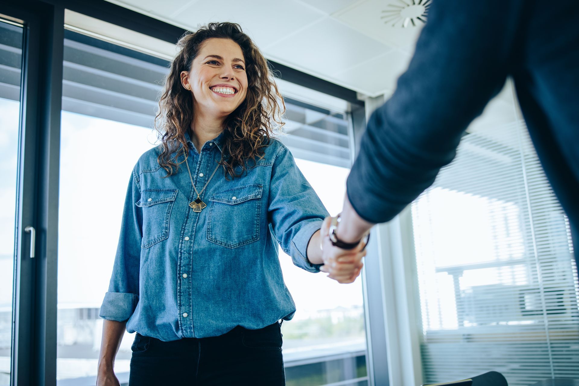 A woman in a denim shirt is shaking hands with a man in an office.