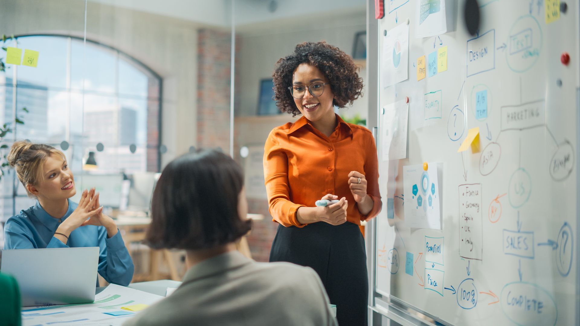 A woman is standing in front of a whiteboard giving a presentation to a group of people.