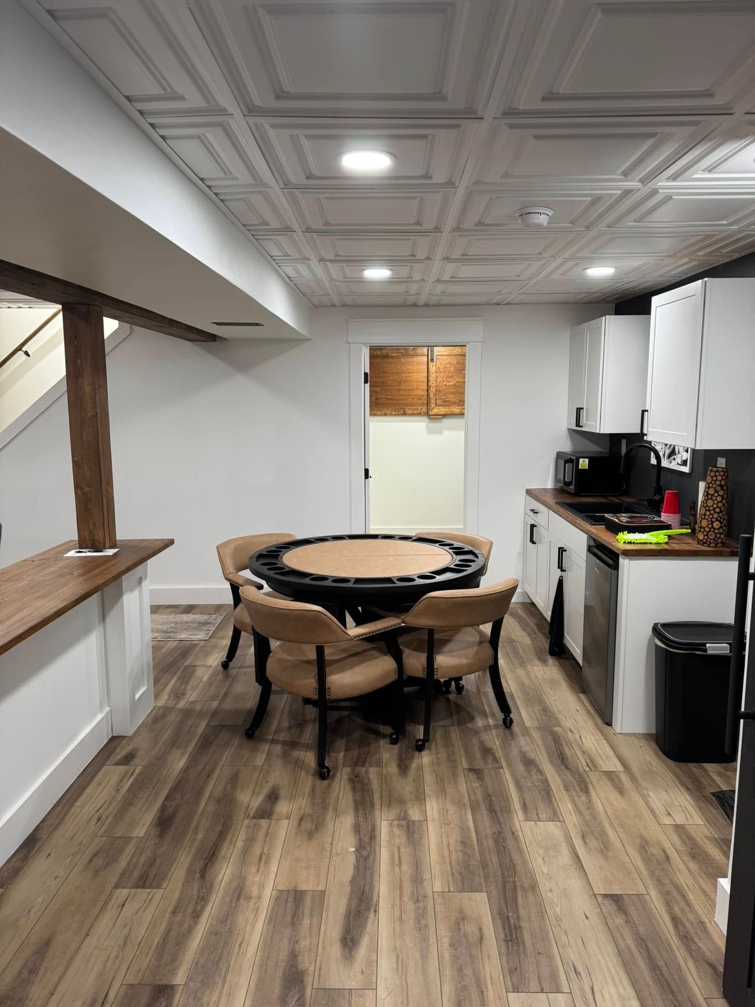 Basement with poker table, chairs, kitchen, and decorative ceiling. Wooden floor and white walls.