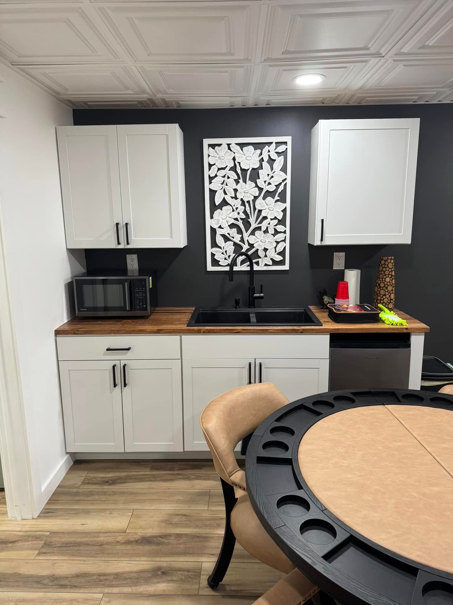 Basement kitchen with white cabinets, wood countertop, black sink, and a decorative black and white wall art.