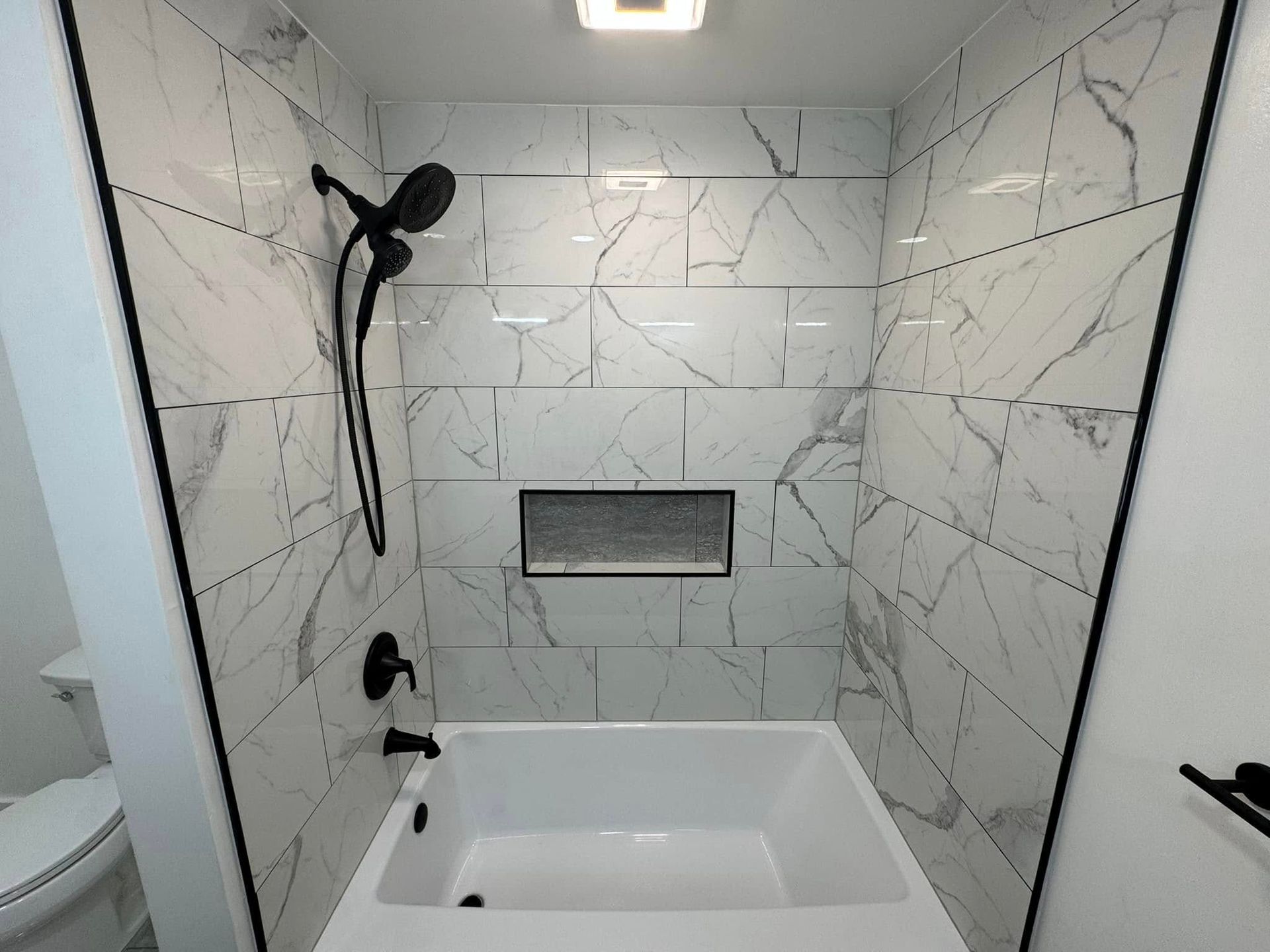 Modern bathroom with marble-look tiled shower and bathtub, black fixtures, and recessed shelf.