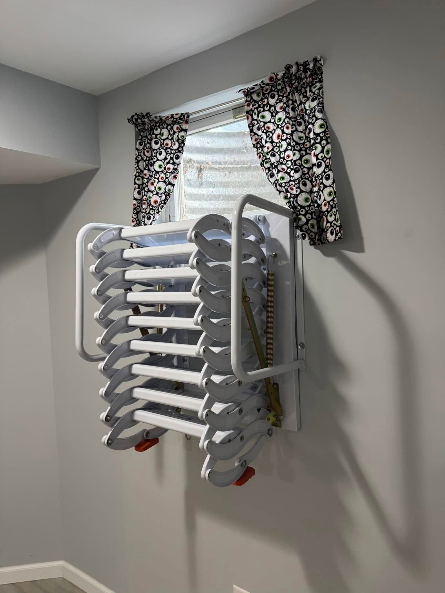 White folding escape ladder mounted under a window with patterned curtains on a gray wall.