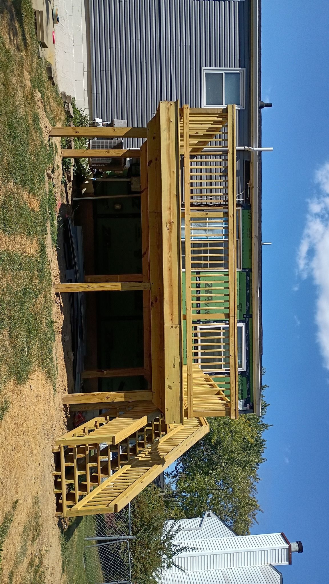 Wooden deck with stairs, built against a building with corrugated siding. Green grass and blue sky.