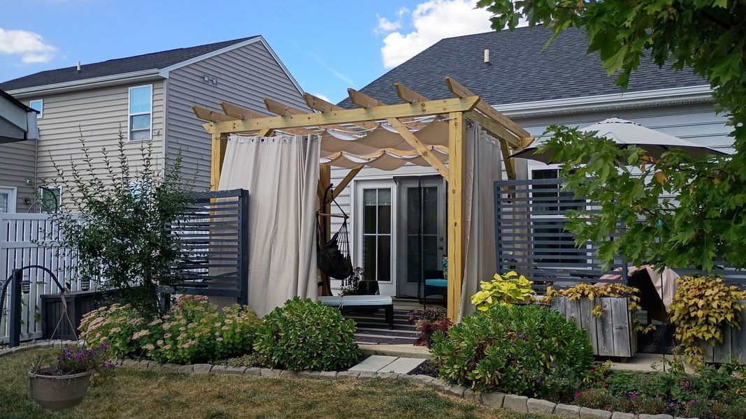 Wooden pergola with tan curtains, in a backyard with landscaping, leading to patio doors.