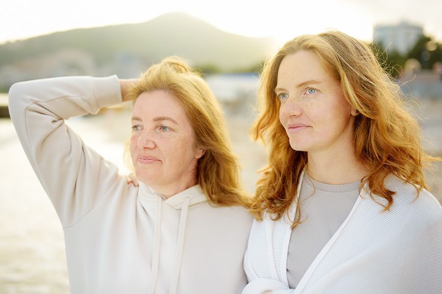 Two women outdoors by water, one with hand on head, looking to the side. Soft sunlight.