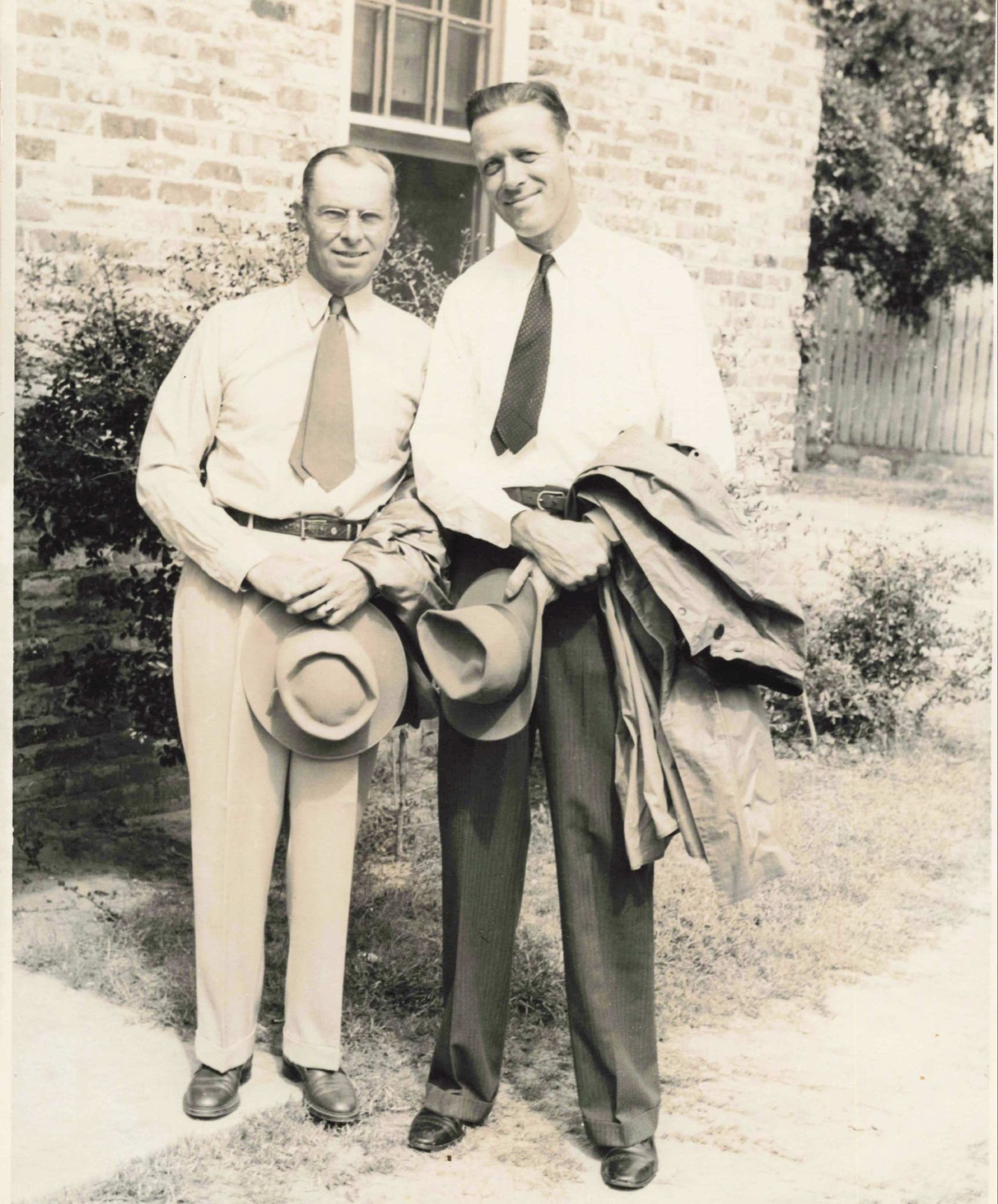 Two men stand outside a brick building. One holds a hat and the other a coat, both wearing shirts and ties.