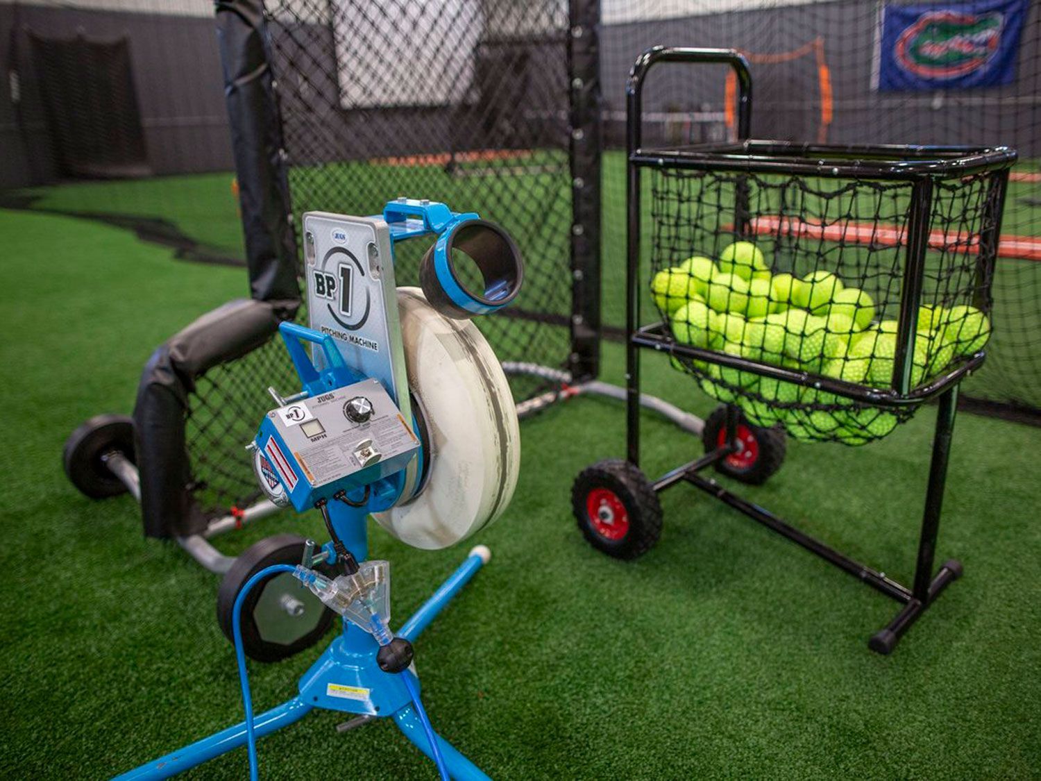 A pitching machine is sitting next to a cart filled with tennis balls.