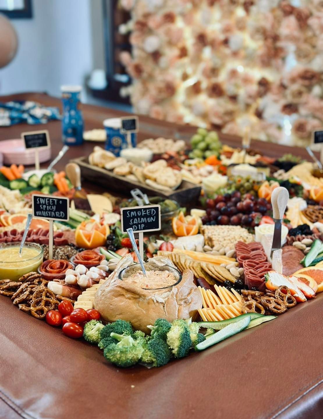 Charcuterie board on a brown surface, filled with cheese, crackers, fruits, vegetables, and dips, with floral backdrop.