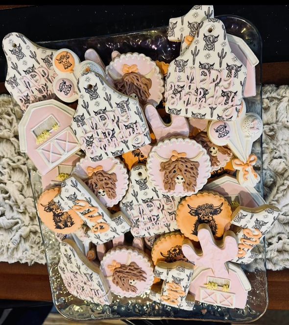 Assortment of decorated sugar cookies on a tray. Includes cow, barn and clothing shapes, with pink, white and brown icing.