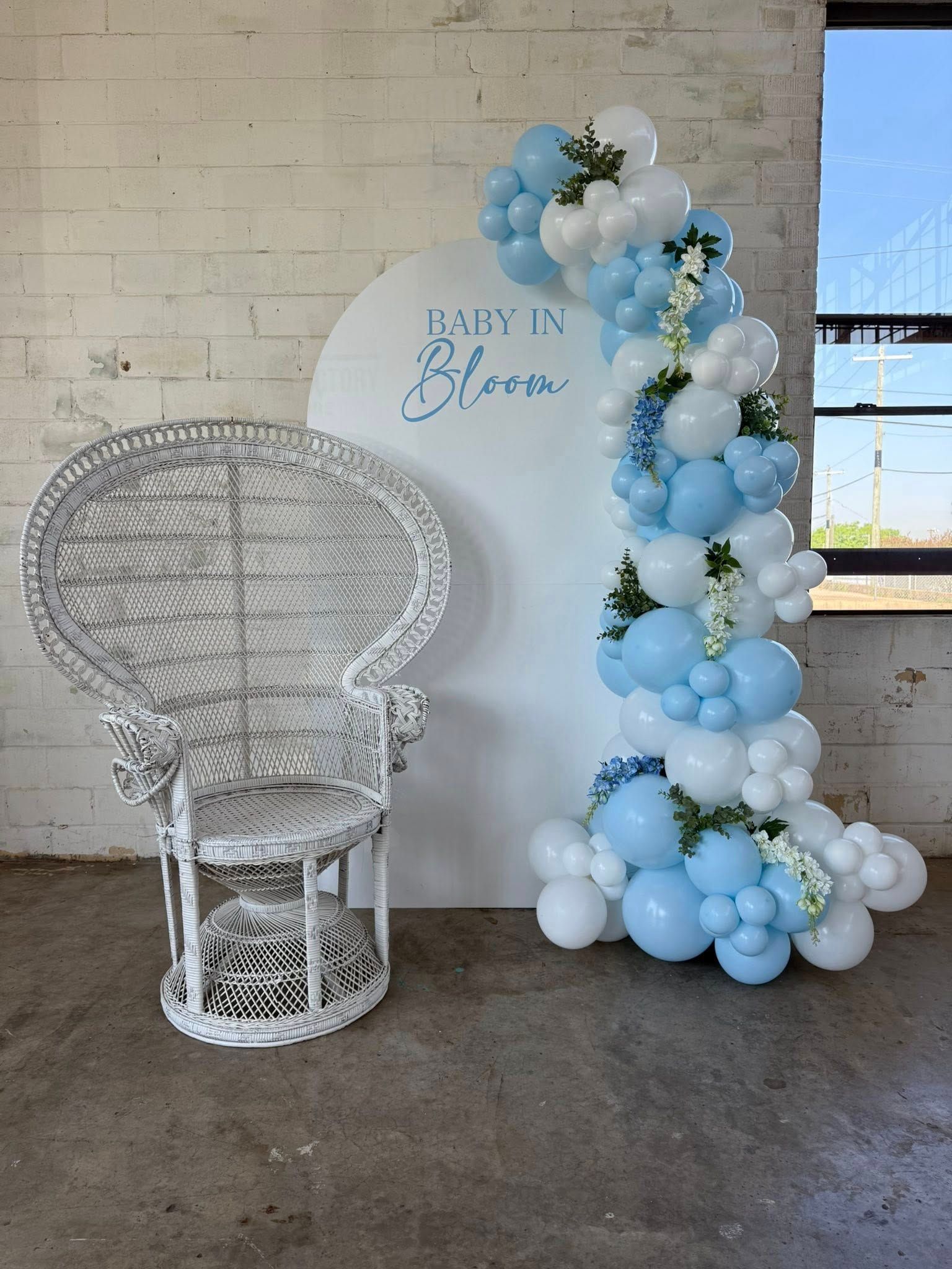White wicker chair, blue and white balloon arch, and a sign that reads