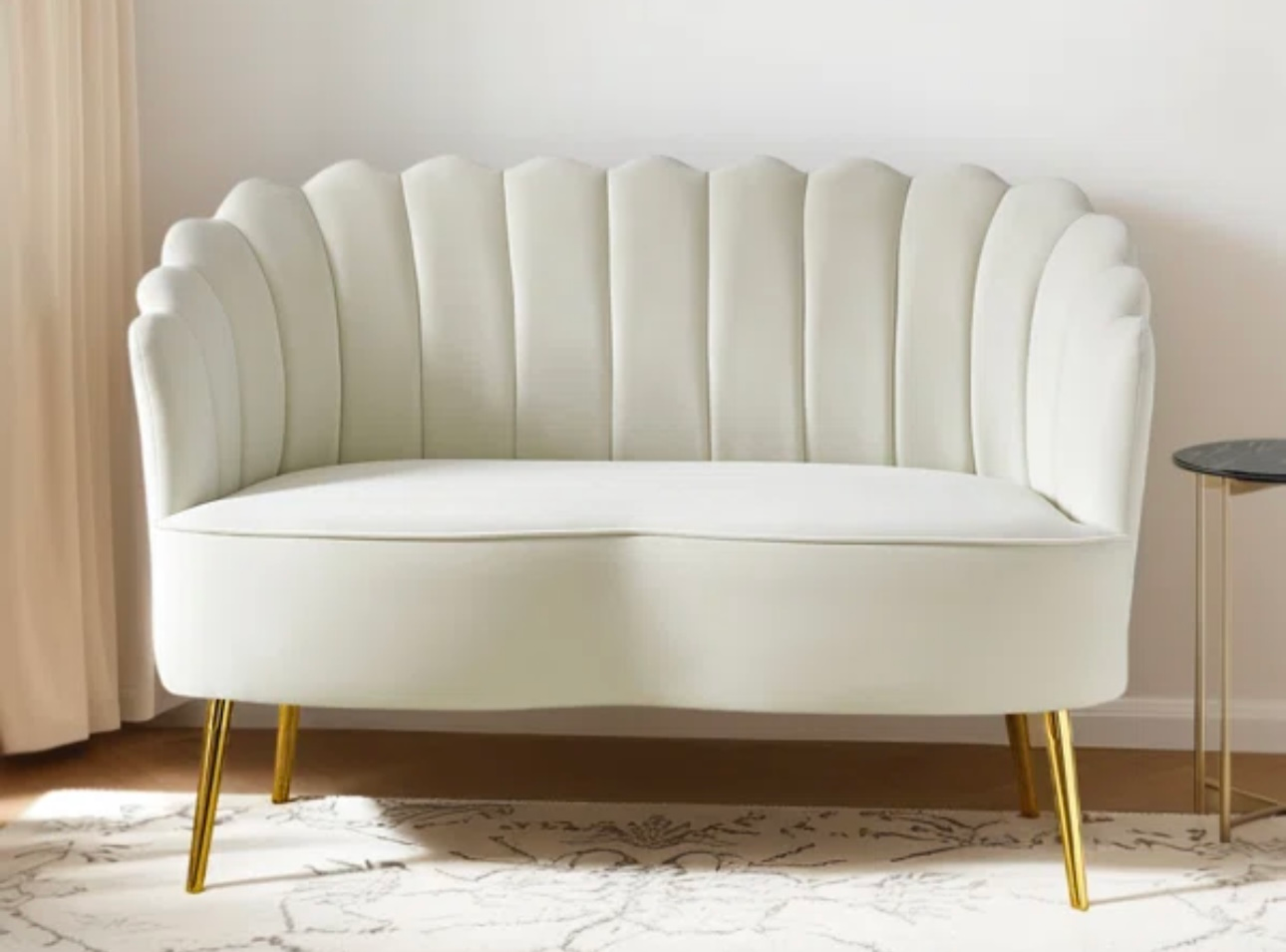Cream-colored loveseat with scalloped back, gold legs, in a room with a side table and rug.