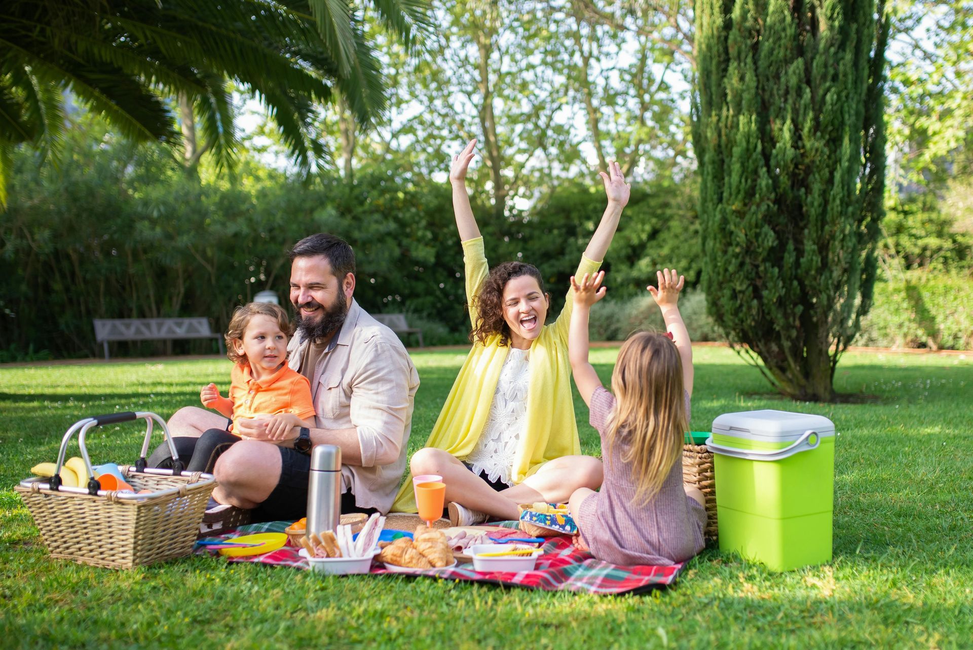 A family of four enjoys a picnic on a sunny lawn, with parents and two young children sitting on a blanket.