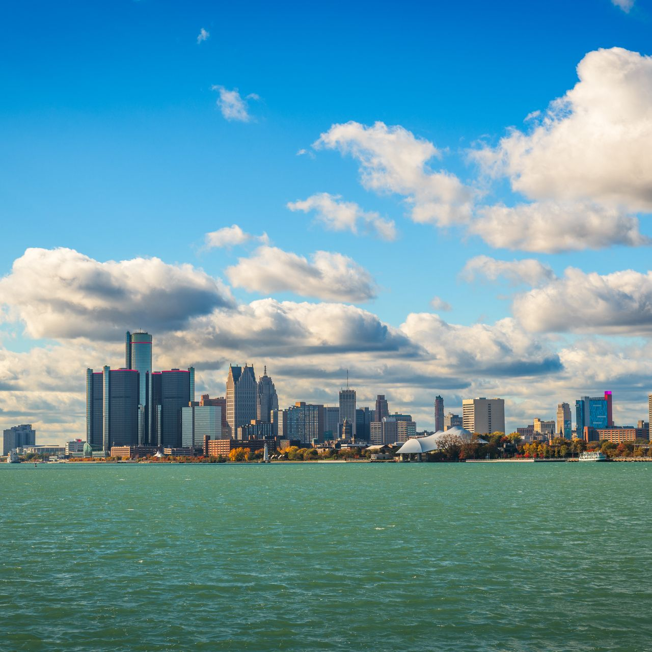 Detroit skyline, tall buildings, against a cloudy blue sky over a green lake.