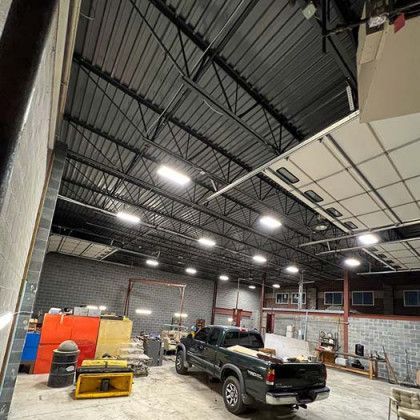 Interior view of a large workshop or garage with a black truck parked in the center. Overhead lighting, brick walls, and a high, angled ceiling.