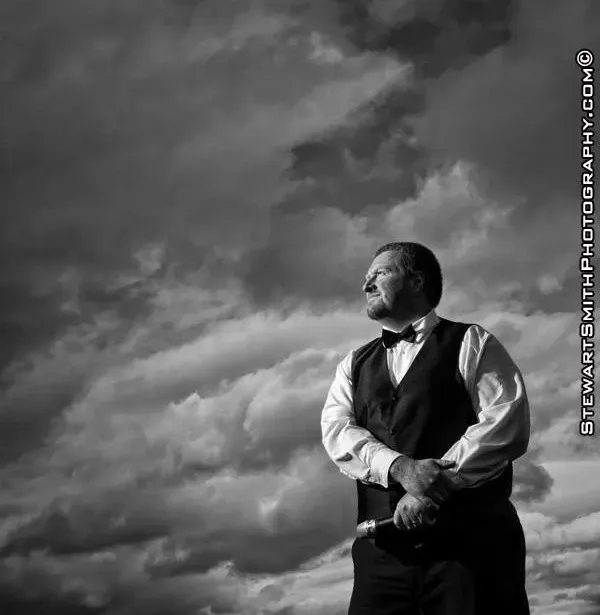 A man in a tuxedo is standing in front of a cloudy sky.