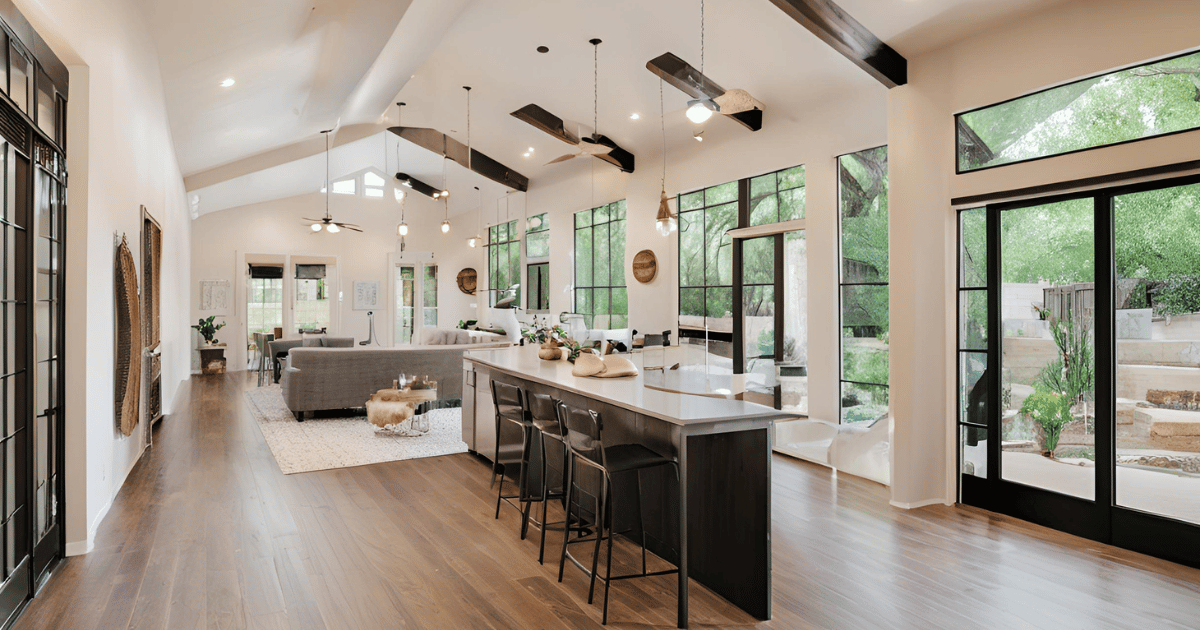 A kitchen with a long island and stools in a large room with lots of windows.