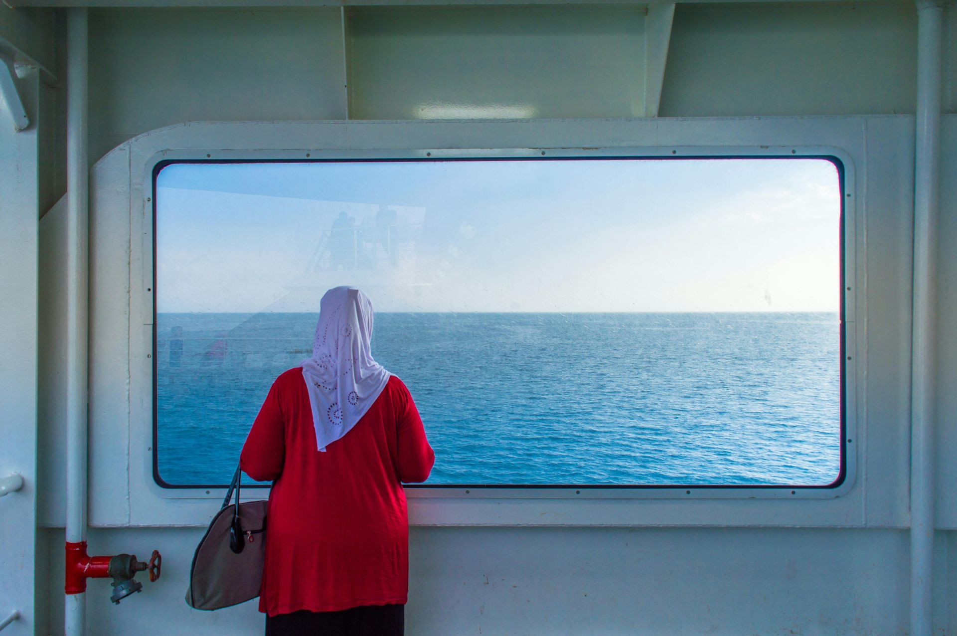 A woman in a hijab is looking out of a window at the ocean.