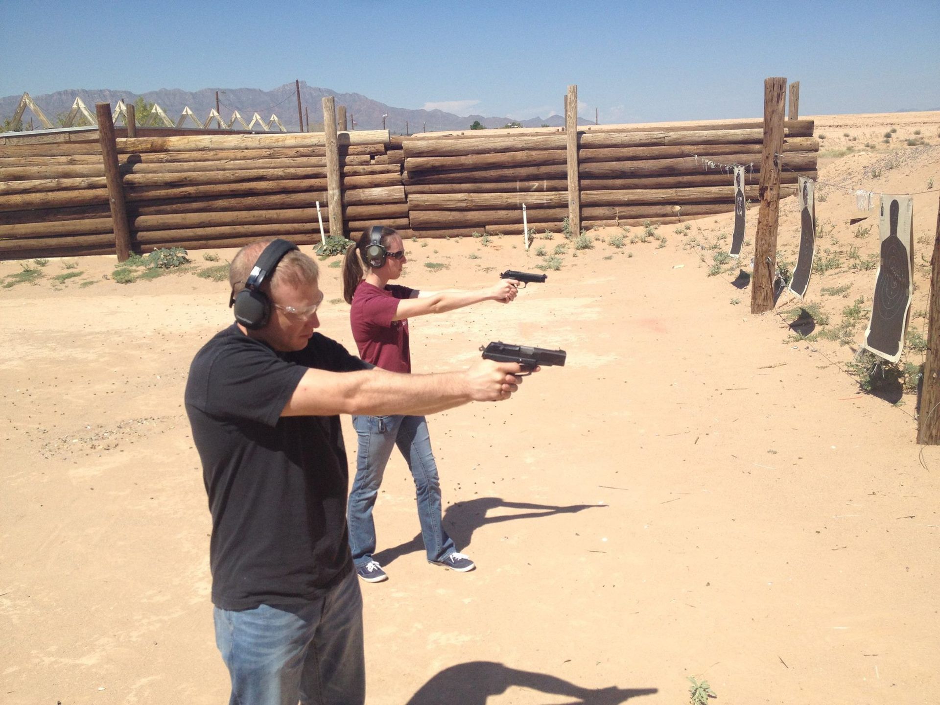 Two people firing handguns at targets at an outdoor shooting range on a sunny day.