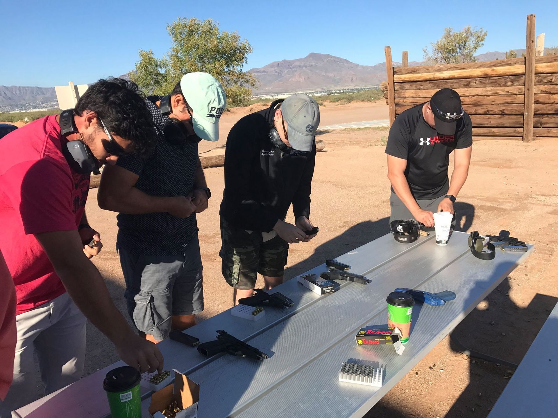 People at a shooting range preparing firearms. Handguns on a table, all wearing ear protection, outdoors.