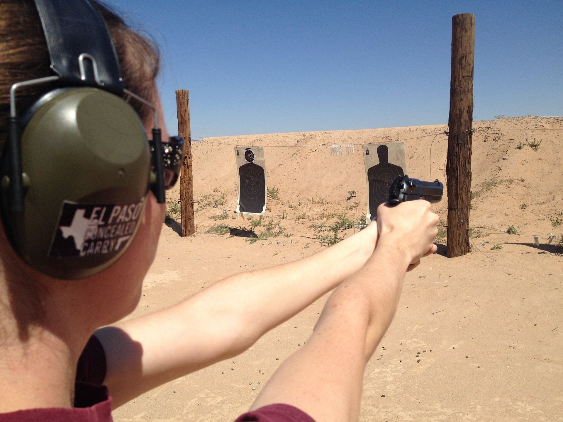 Person aiming a handgun at a target on a shooting range, wearing ear protection. Sunny day.