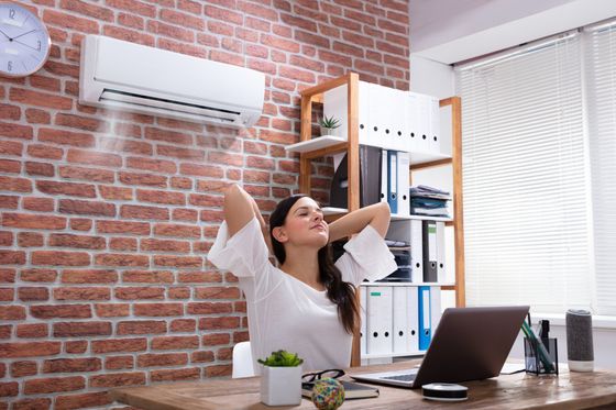 A woman is sitting at a desk with her arms outstretched in front of an air conditioner.