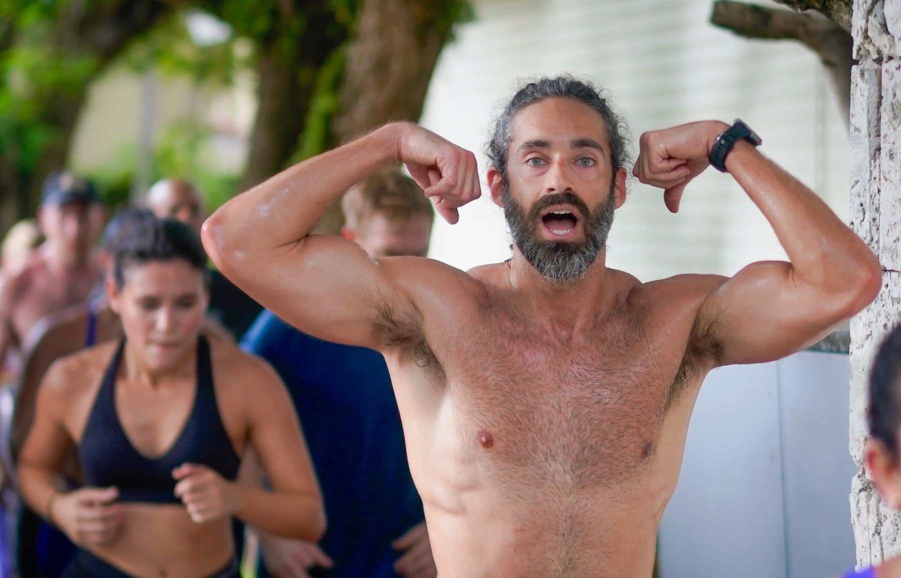 A shirtless man with a beard is flexing his muscles in front of a group of people.