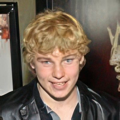 A young man with blonde hair and blue eyes is wearing a black jacket