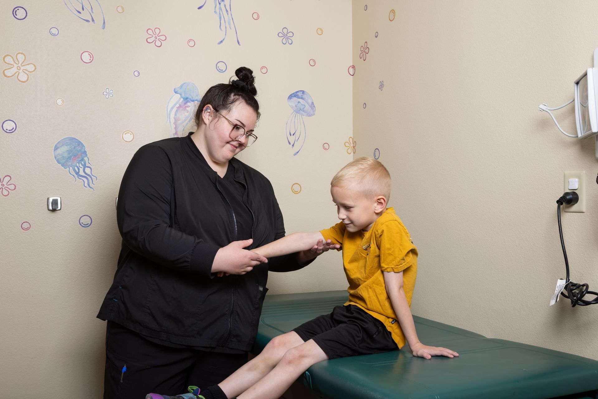 A child receives medical attention in a clinic. A doctor and parent assist; colorful kids' art decorates the wall.